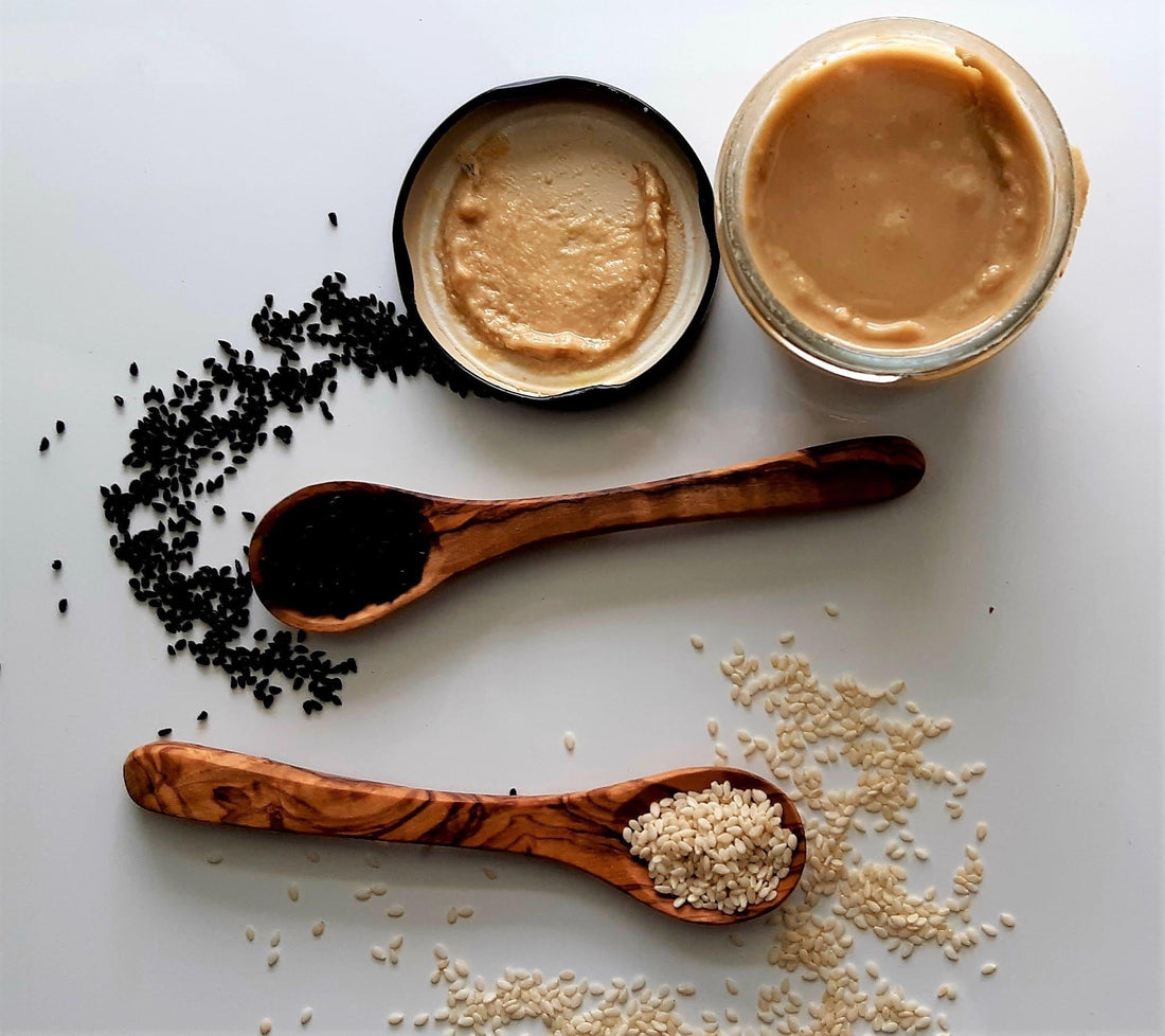     Black and white sesame seeds arranged with wooden spoons and bowls of seed butter on a neutral background.